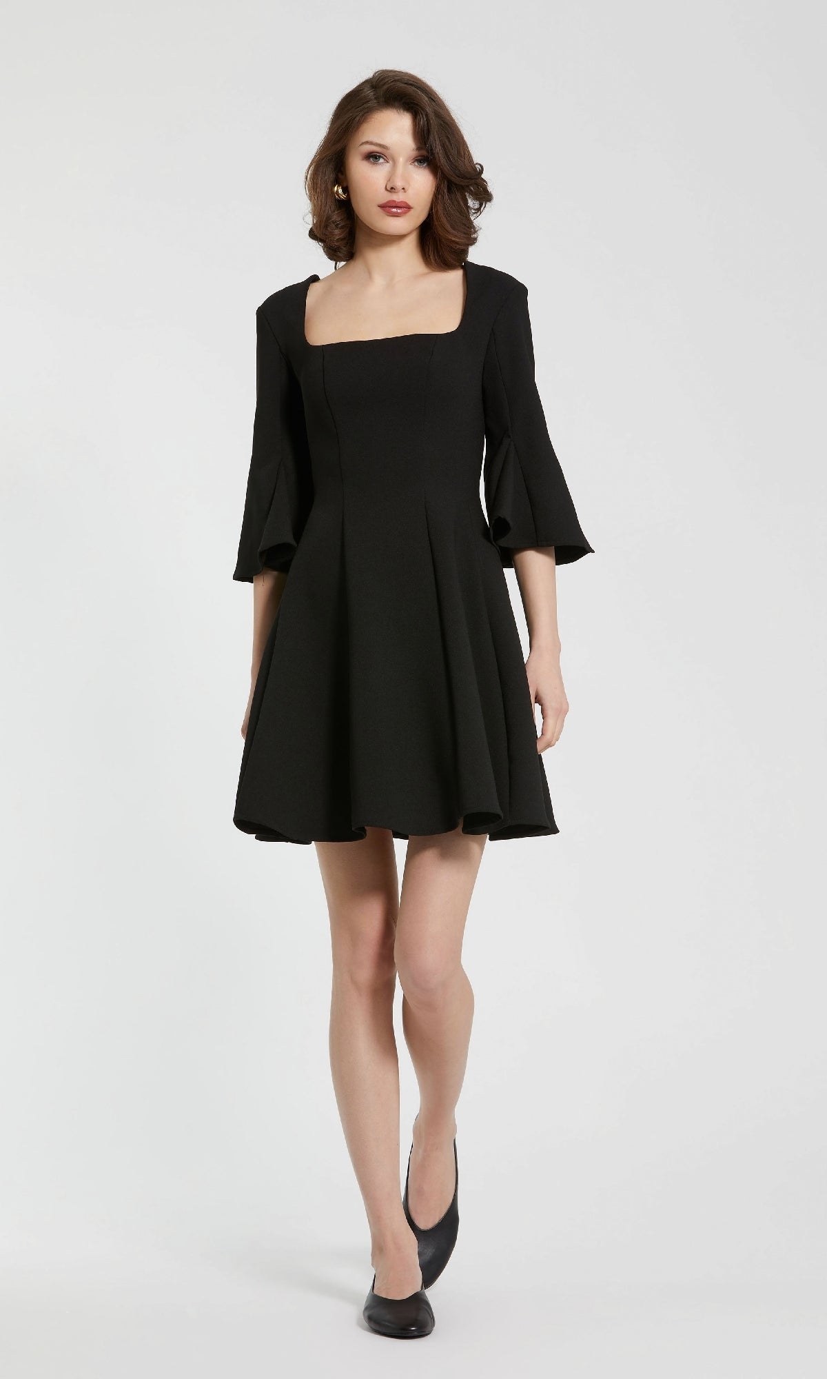 A woman in the Mac Duggal 21011 Cute Short Black Party Dress with flared sleeves and black flats stands against a plain white background.